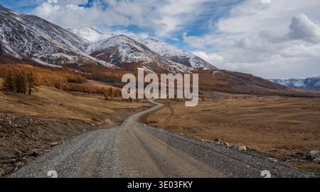 Unbefestigte Straße, die sich durch die wunderschöne Herbstlandschaft mit verschneiten Bergen und spärlich verstreuten Bäumen schlängelt, atemberaubende Naturlandschaft Stockfoto