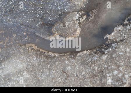 Schmelzendes Schneebad auf Asphaltoberfläche, schmutziger Schlamm bildet einen schmalen braunen Kanal, glitzernde Eiskristalle fangen Sonnenlicht ein, Nahaufnahme-Textur-Highlight Stockfoto