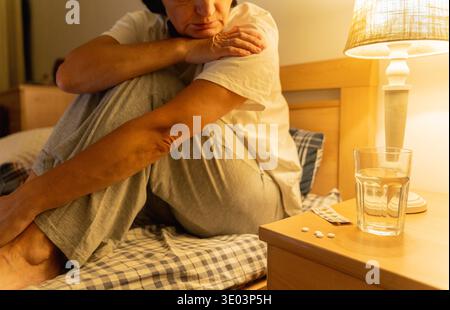 Eine erwachsene Frau, die an Schlaflosigkeit leidet, sitzt verärgert im Bett und ist dabei, Medikamente, Pillen und ein Glas Wasser auf dem Nachttisch zu nehmen Stockfoto