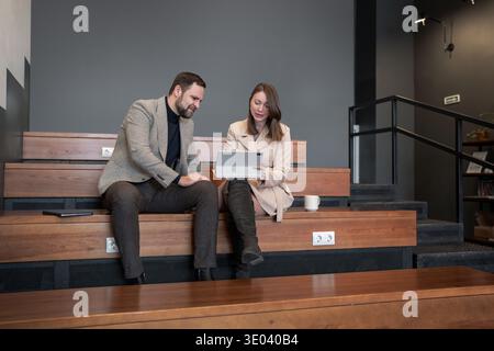 Geschäftsmann und Geschäftsfrau, die in stilvollem Büro auf Holztreppen auf Laptop zeigen. Zusammenarbeit auf Notebooks, Teamarbeit, Büro, Planung Stockfoto