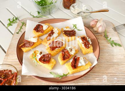 Heiße, knusprige Polenta-Stücke, gebacken mit cremigem Blauschimmelkäse und sonnengetrockneten Tomaten, serviert auf Terrakotta-Teller mit Rucola, Knoblauch und Serviette Stockfoto