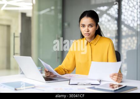 Junge Geschäftsfrau, die in die Kamera schaut, verschiedene Dokumente hält und an ihrem Schreibtisch in einem modernen Firmenbüro an einem Laptop arbeitet, wobei Fokus und Engagement hervorgehoben werden Stockfoto