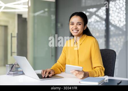 Lächelnde junge Geschäftsfrau, die fleißig in einem modernen Büro arbeitet, einen Laptop und ein digitales Tablet verwendet und Technologie, Professionalität und Erfolg in einer kollaborativen Unternehmensumgebung verkörpert Stockfoto