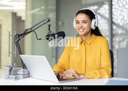 Weibliche Podcaster, die Kopfhörer trägt und die Kamera anlächelt, während sie auf einem Laptop schreibt und neues Audiomaterial aus einem modernen Studio aufnimmt und sendet Stockfoto