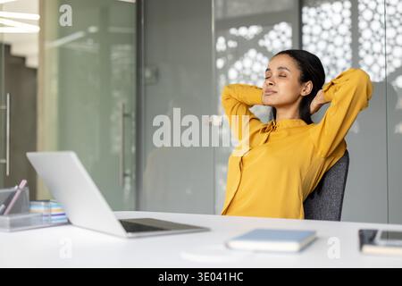 Frau, die einen achtsamen Moment nimmt, entspannt sich mit geschlossenen Augen und Händen hinter dem Kopf und genießt während einer Pause an ihrem modernen Büroarbeitsplatz Stressabbau Stockfoto