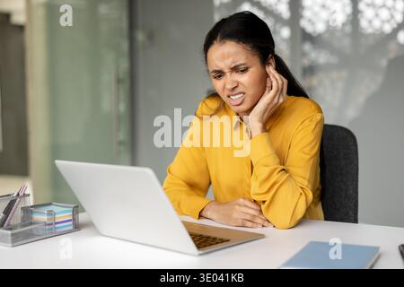 Eine junge Geschäftsfrau, die an schweren Ohrenschmerzen und einer Ohrentzündung leidet und ihr schmerzhaftes Ohr berührt, während sie sich mit einem Laptop auf ihre Arbeit an einem modernen Schreibtisch konzentriert Stockfoto