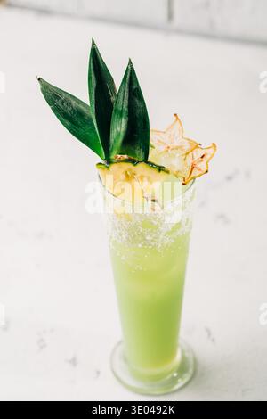 Lebendiger tropischer Cocktail in einem hohen Glas, garniert mit frischen Ananasblättern und Sternenfrüchten, auf einer Marmorfläche. Perfekt zum Zusammenziehen im Sommer Stockfoto