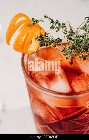 Eine Nahaufnahme eines erfrischenden Cocktails in einem gravierten Glas mit einem orangen Twist und Kräutern. Das Getränk sitzt auf einer Marmoroberfläche, Eis schmilzt Stockfoto