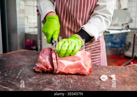 Ein erfahrener Metzger schneidet mit einer gestreiften Schürze und hellgrünen Handschuhen auf einem Holzschneidebrett rohes Fleisch in einem gut ausgestatteten Profi Stockfoto