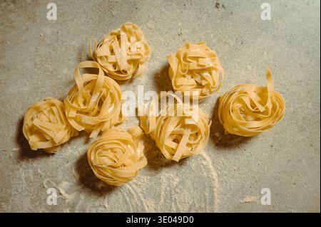 Ein Blick von oben auf frisch zubereitete Tagliatelle Nudelnester, die auf einer bemehlten Oberfläche angeordnet sind. Der leuchtende gelbe Farbton der ungekochten Nudeln unterstreicht die Frische und die Frische Stockfoto