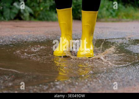 Ein Paar hellgelbe Stiefel platzen in einer schlammigen Pfütze auf einem unbefestigten Pfad, umgeben von üppigem Grün, und symbolisieren einen abenteuerlichen Geist während eines regnerischen Tages Stockfoto