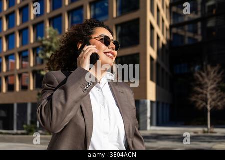 Selbstbewusste Geschäftsfrau mit lockigem Haar, die eine Sonnenbrille trägt, lächelt, während sie mit ihrem Smartphone in einer modernen städtischen Umgebung spricht, verkörpert Profi Stockfoto