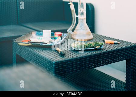 Eine Nahaufnahme eines Korbtisches mit Cannabisknospen auf einem Tablett, einem gerollten Joint und verschiedenen Raucheraccessoires, die vor legeren Sitzgelegenheiten im Freien stehen Stockfoto