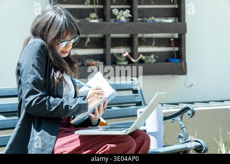 Frau, die im Freien arbeitet, telefoniert und in einem Notebook mit Laptop an ihrer Seite schreibt Stockfoto