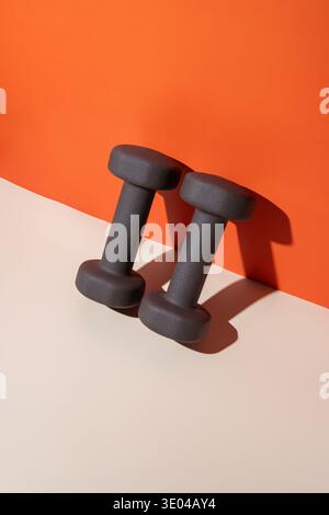 Graue Kurzhanteln auf beigefarbener Oberfläche mit auffälligem orangefarbenem Hintergrund, ideal für Krafttraining- und Fitnessbegeisterte, die nach Enha suchen Stockfoto