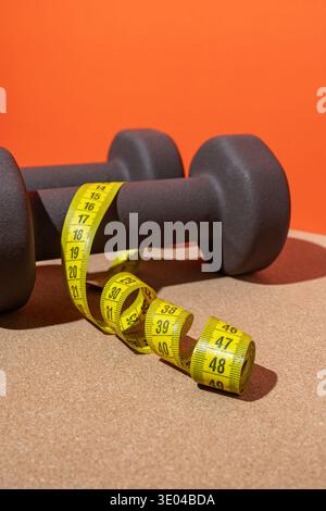 Zwei Kurzhanteln mit gelbem Maßband auf Korkoberfläche, vor einem leuchtend orangen Hintergrund, die Fitness, Bewegung und gesundes Leben symbolisieren Stockfoto
