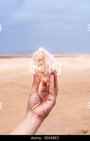 Eine Hand, die einen rohen Salzsteinkristall vor dem Hintergrund der Skeleton Coast in Namibia hält. Der Kristall zeigt komplizierte natürliche Formationen darunter Stockfoto