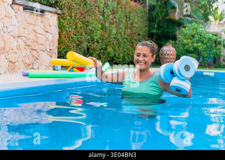 Eine Frau in einem Swimmingpool lächelt beim Training mit bunten Schaumnudeln und Kurzhanteln. Der Poolbereich bietet üppiges Grün und einen Stein Stockfoto