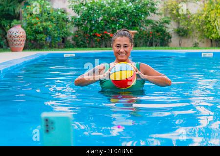 Fröhliche Frau in grünem Badeanzug, die einen entspannenden Tag im Pool mit einem lebhaften Strandball genießt. Umgeben von üppigen Pflanzen fängt die Szene Su ein Stockfoto