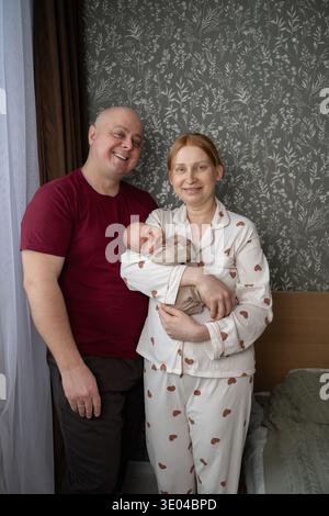 Ein fröhlicher Moment zu Hause mit einer lächelnden Familie, die die Ankunft ihres Neugeborenen feiert. Die Eltern sind in kuscheliger Kleidung gekleidet Stockfoto