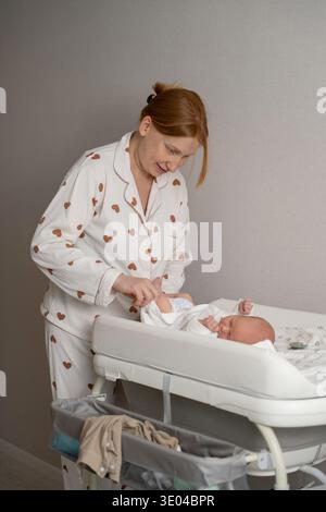 Eine Mutter wechselt liebevoll die Windel ihres Neugeborenen zu Hause. Sie trägt einen Pyjama mit Herzmuster, lächelt zärtlich und schafft eine warme, fürsorgliche Atmosphäre Stockfoto