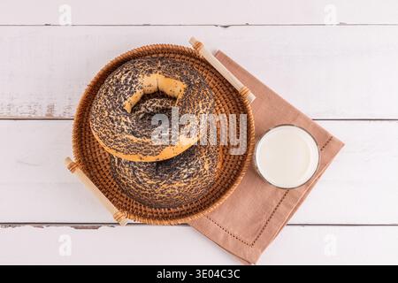 Zwei Mohnsäcke ruhen auf einem Korbteller neben einem Glas Milch auf einer braunen Serviette. Der darunter liegende Holztisch ergänzt die warmen Töne des Stockfoto