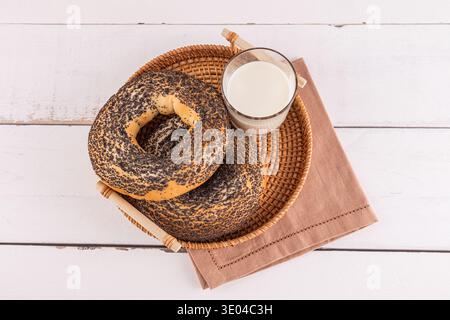 Zwei frisch gebackene Vollkornbagels, die mit Mohn bedeckt sind, ruhen in einem Webkorb neben einem Glas Milch auf einer braunen Serviette über einem rustikalen W Stockfoto
