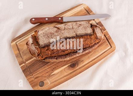 Ein frisch gebackener Vollkornbrot liegt auf einem hölzernen Schneidebrett und zeigt seine rustikale Textur. Ein gezacktes Messer liegt in der Nähe, bereit zum Schneiden Stockfoto