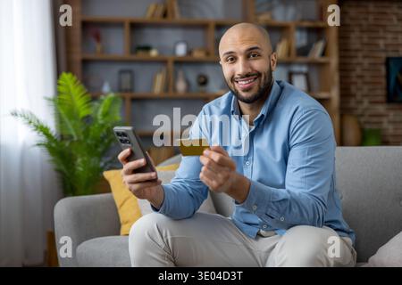 Mann bezahlt online mit einer Kreditkarte, hält ein Smartphone und lächelt in die Kamera, während er bequem auf einem Sofa zu Hause sitzt, genießt bequem Stockfoto