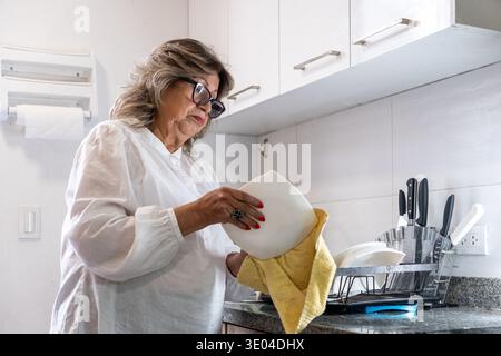 Eine ältere Frau, die nach dem Waschen eine saubere weiße Schüssel mit einem gelben Handtuch abwischt Stockfoto