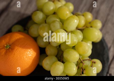 Nahaufnahme einer sonnendurchfluteten rustikalen Tischplatte mit einer engen Gruppe grüner kernloser Trauben neben einer leuchtenden Orange auf einer dunklen Platte, die glänzende Häute hervorhebt; Stockfoto