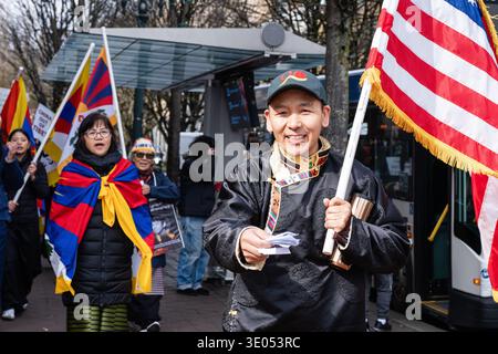 Portland, Oregon, USA. März 2026. Ein Mann in traditioneller tibetischer Kleidung trägt eine US-Flagge und hält gedruckte Materialien, während sich die Teilnehmer zu einem von der Northwest Tibetan Cultural Association organisierten Tibetischen Aufstand-marsch versammeln. Eine Frau mit einer tibetischen Flagge geht hinter ihm her. Quelle: Norboe Photo/Alamy Live News Stockfoto