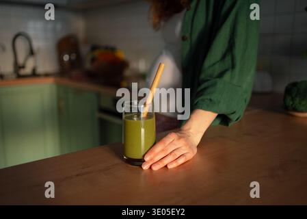 Schwangere Frau Hand auf einer Theke, während sie in der Nähe eines Glases grünen Smoothies mit Bambusstroh steht und sich auf gesunde Ernährung und Selbstpflege in ihrer Küche konzentriert Stockfoto