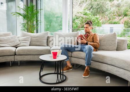 Mann sitzt direkt auf geschwungenem Sofa im Wohnzimmer und hält Fußball, Tasse, Popcorn, Kopierraum. Erwachsene, Ball, konzentriert, Smartwatch, modern, Innenraum, Heim Stockfoto
