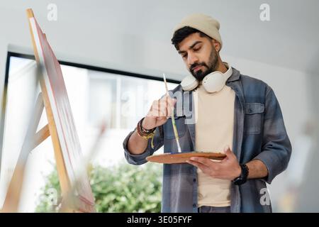 Indischer Mann malte an der Staffelei mit Palette und Pinsel, trug Beanie und Headset am Studiofenster. Künstler, Tageslicht, Leinwand, minimalistisch, Natur, foc Stockfoto