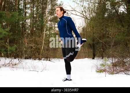 Männlicher Läufer, der in einem schneebedeckten Wald eine Quad-Strecke macht. Aktiver junger Mann, der sich für ein Outdoor-Workout im Winter in sportlicher Sportbekleidung aufwärmt. Hochwertiges Foto Stockfoto