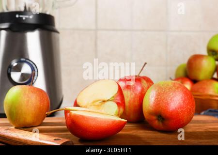 Frische rote Äpfel, ganz und in Scheiben geschnitten, auf einem hölzernen Schneidebrett mit Küchenmesser, neben einem Edelstahl-Mixer. Eine gesunde Lebensmittelzubereitung Scen Stockfoto