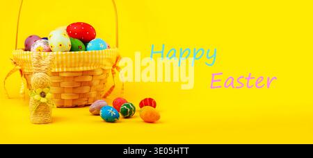 Ein fröhliches Osterbanner mit einem Korbkorb gefüllt mit handbemalten Eiern, einem Strohhasen und einem „Happy Easter“-Text auf einer leuchtenden gelben Rückseite Stockfoto