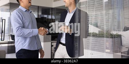 Zwei multiethnische Geschäftsleute halten ein digitales Tablet und schütteln sich im Büro die Hände Stockfoto