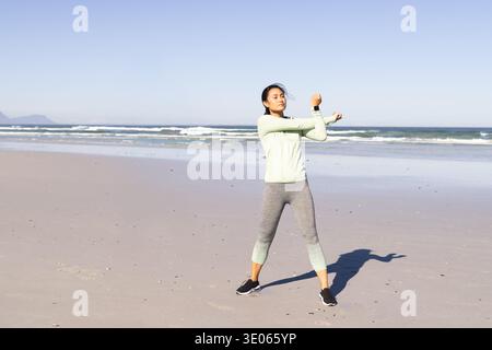 Asiatische Frau, die Arm dehnt am Sandstrand in der Nähe der Küste in grüner Spitze, capri-Leggings, Uhr. Am Meer, im Freien, Fitness, Stretching, Wellen, Ozean, Stockfoto
