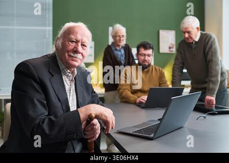 Ein älterer kaukasischer Mann sitzt am Tisch und hält einen Stock, während eine ältere kaukasische Frau, ein älterer kaukasischer Mann und ein junger erwachsener kaukasischer Mann an einem Computerkurs mit Laptops teilnehmen Stockfoto
