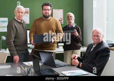 Gruppe von älteren kaukasischen Männern und Frauen mit einem jungen Erwachsenen kaukasischen Mann, der an einem Computerkurs teilnimmt, Laptops hält und mit offenen Notebooks und Geräten am Tisch sitzt Stockfoto