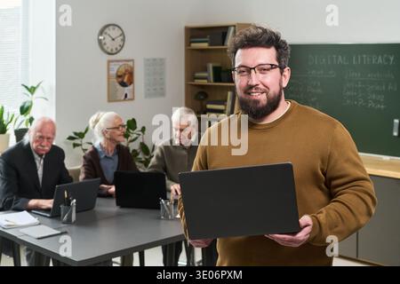 Porträt eines kaukasischen jungen Erwachsenen, der einen Laptop hält und in die Kamera lächelt, während ältere kaukasische Männer und Frauen während des Computerkurses mit Laptops am Tisch sitzen Stockfoto