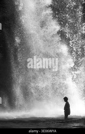 Kleines Kind, das im Wasser steht, während der mächtige Wasserfall herumspritzt Stockfoto
