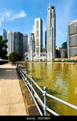 Die Leute laufen entlang des Kanals in Singapur. Hohe Gebäude ragen auf beiden Seiten unter klarem Himmel auf. Bäume verleihen der Szene einen Hauch von Grün. Stockfoto