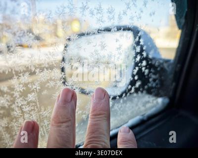 Frost bildet komplizierte Muster auf dem Autofenster, während die Person es an einem kalten Wintermorgen in der Nähe einer geschäftigen Straße berührt. Stockfoto