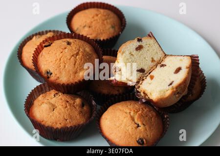 Platte mit hausgemachten Rosinenmuffins, gebacken in braunen Papiereinlagen, wobei ein aufgeschnittener Muffin weiche Schwammbrösel und Rosinen zeigt Stockfoto