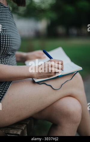 Frau, die mit einem blauen Stift und überkreuzten Beinen in Notizbuch auf einer Parkbank schreibt Stockfoto
