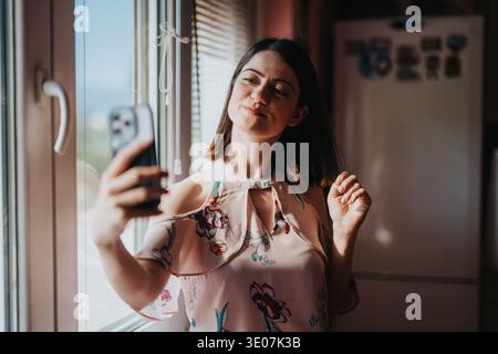 Junge Frau, die zu Hause ein Selfie am Fenster macht, lächelt und posiert mit dem Smartphone Stockfoto