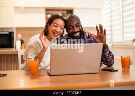 Ein Mann und eine Frau sitzen nebeneinander am Tisch und lächeln warm, während sie einen Videoanruf auf einem Laptop in einem modernen Zuhause führen. Stockfoto